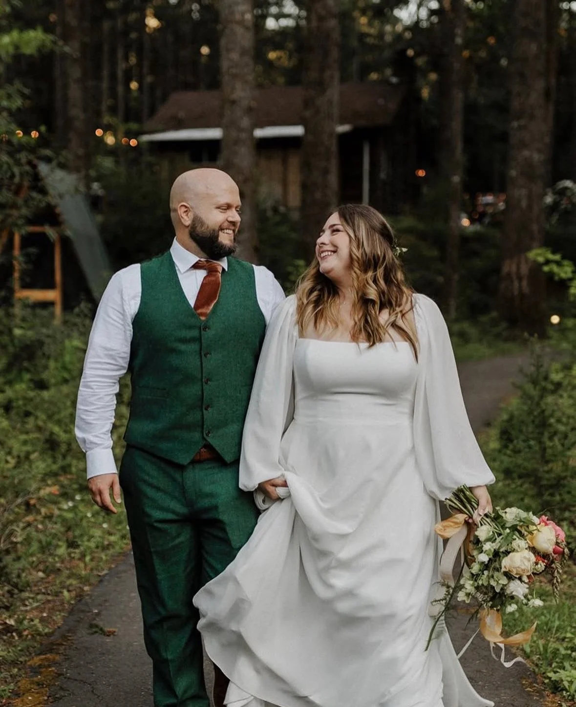 A couple walking outdoors on a wooded path, smiling at each other. The woman wears a white wedding dress and holds a bouquet, while the man wears a green vest, white shirt, brown tie, and green pants.