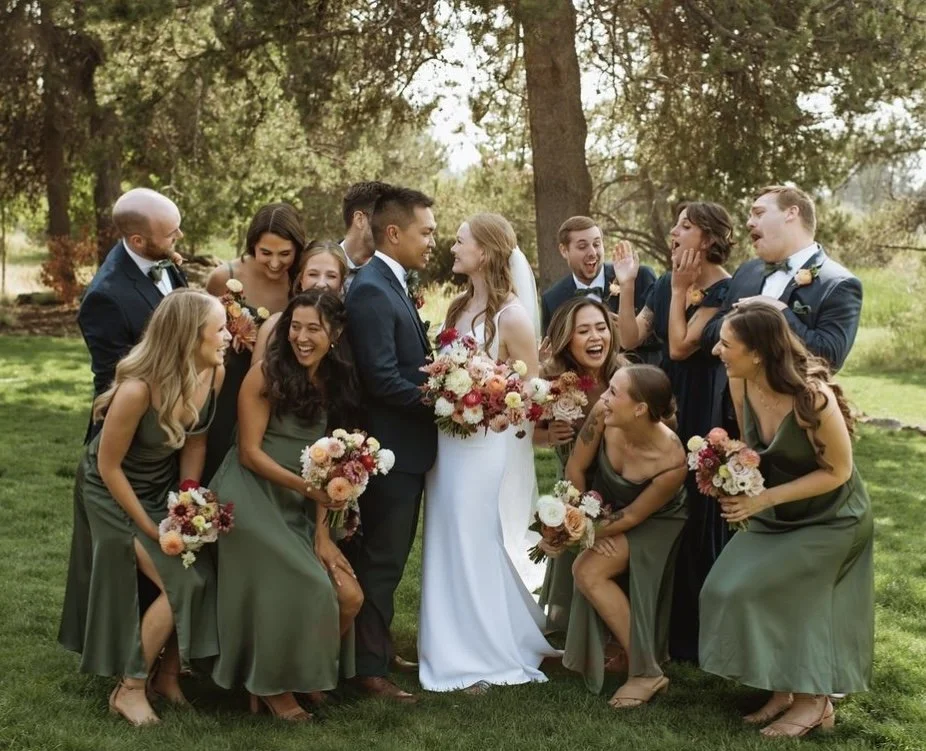 A wedding party of ten people, including the bride and groom, outdoors on a grassy area with trees, dressed in formal attire and holding bouquets, smiling and celebrating.