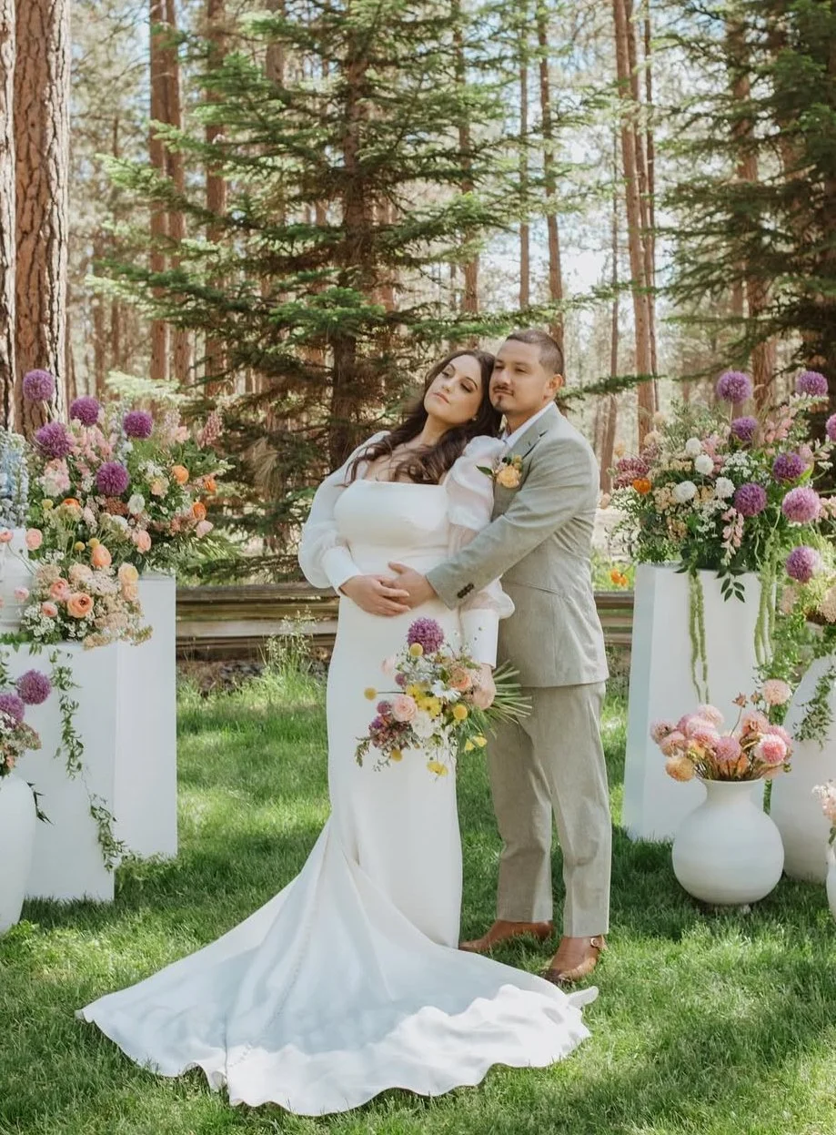 A bride and groom standing among floral arrangements in a forested outdoor setting. The bride wears a white wedding dress, and the groom wears a gray suit. They embrace each other with peaceful expressions.