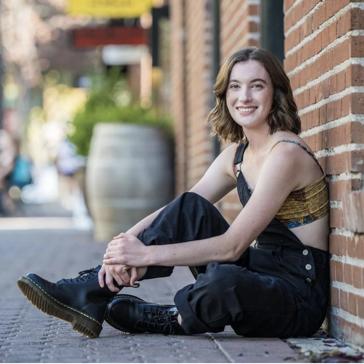 A young woman with shoulder-length wavy brown hair sitting on a sidewalk against a brick wall, smiling, wearing a colorful crop top, black pants, and black combat boots.