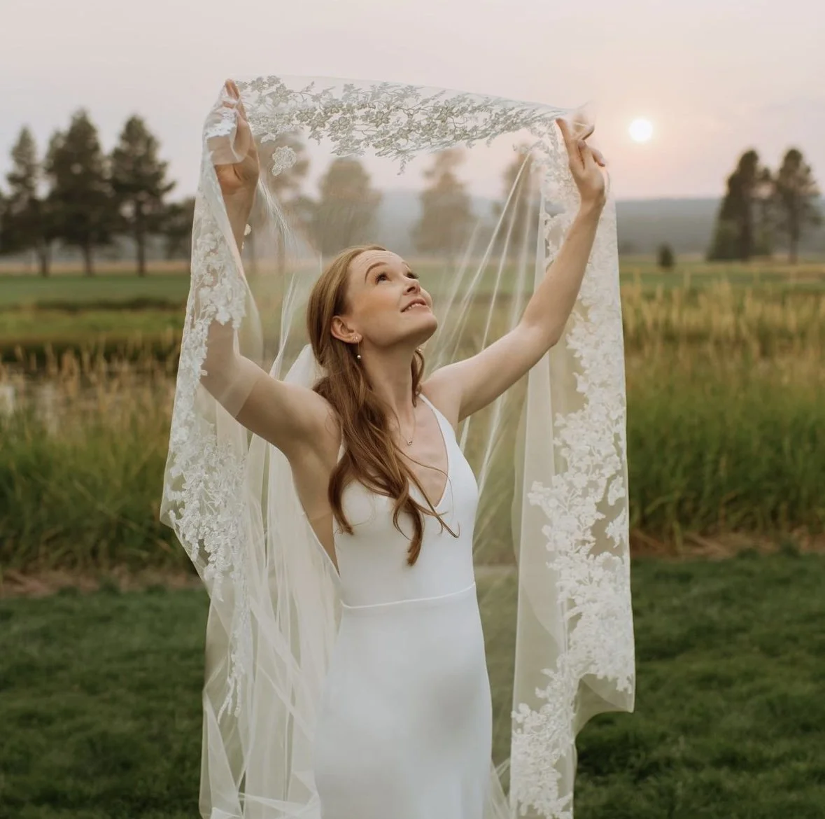 A woman in a white dress holds a lace veil up in the air outdoors during sunset in a grassy field with trees in the background.