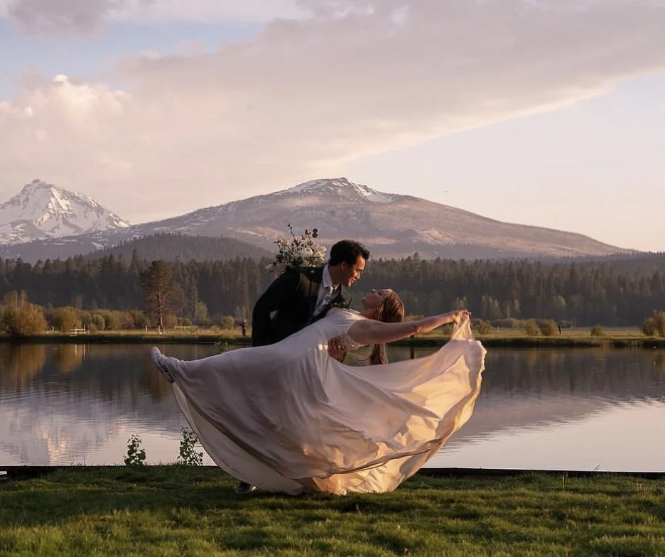 A couple dancing by a lake with mountains in the background during sunset, the woman in a flowing dress and the man in a suit.