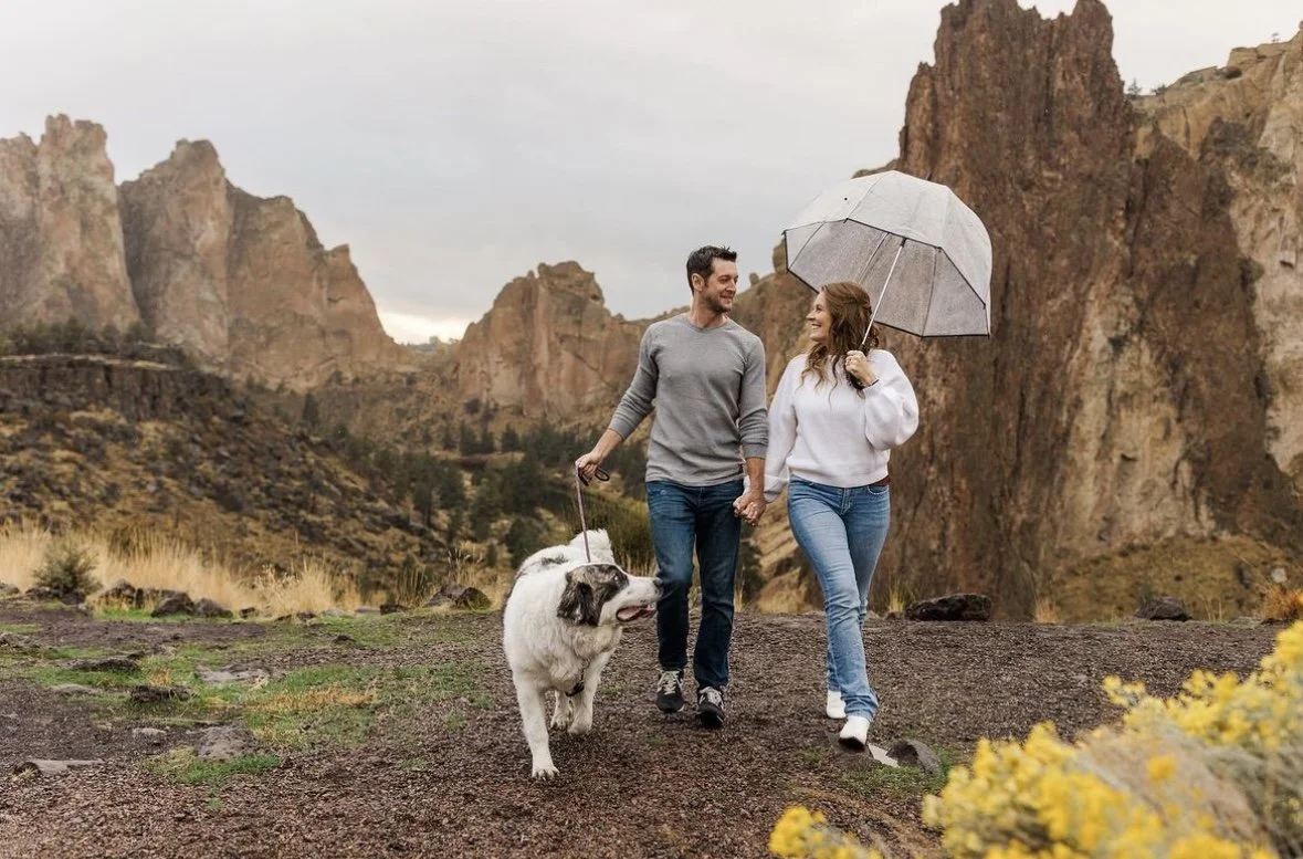 A couple walks holding hands with their dog in a mountainous outdoor landscape, sharing a smile. The woman holds an umbrella, and the sky appears overcast, suggesting rain.