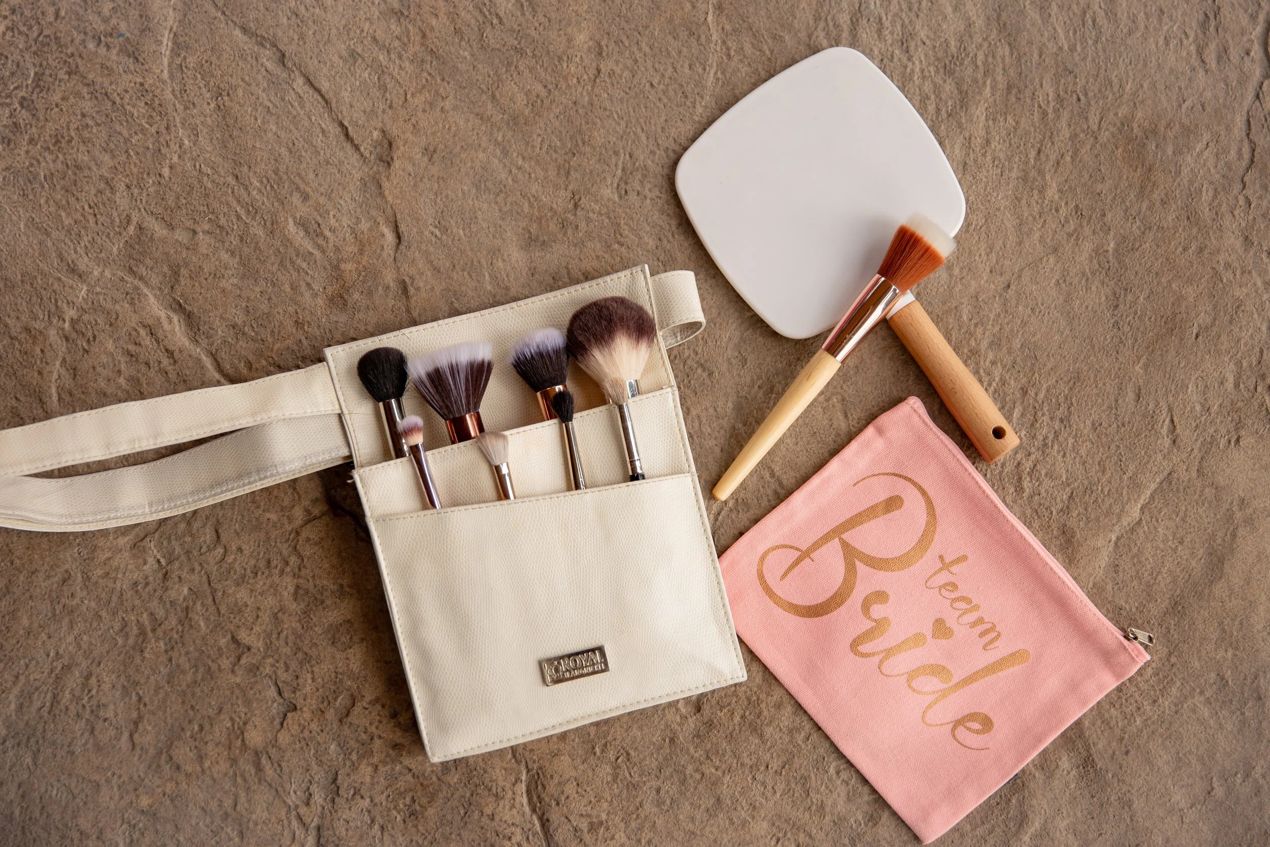 A collection of makeup brushes in a cream-colored roll-up holder, a white square makeup sponge, a makeup brush with a wooden handle, and a pink cloth with gold lettering on a textured brown surface.