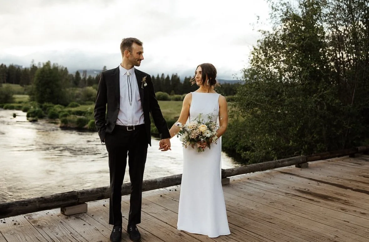A bride and groom holding hands on a wooden bridge next to a river, with greenery and trees in the background under a cloudy sky.