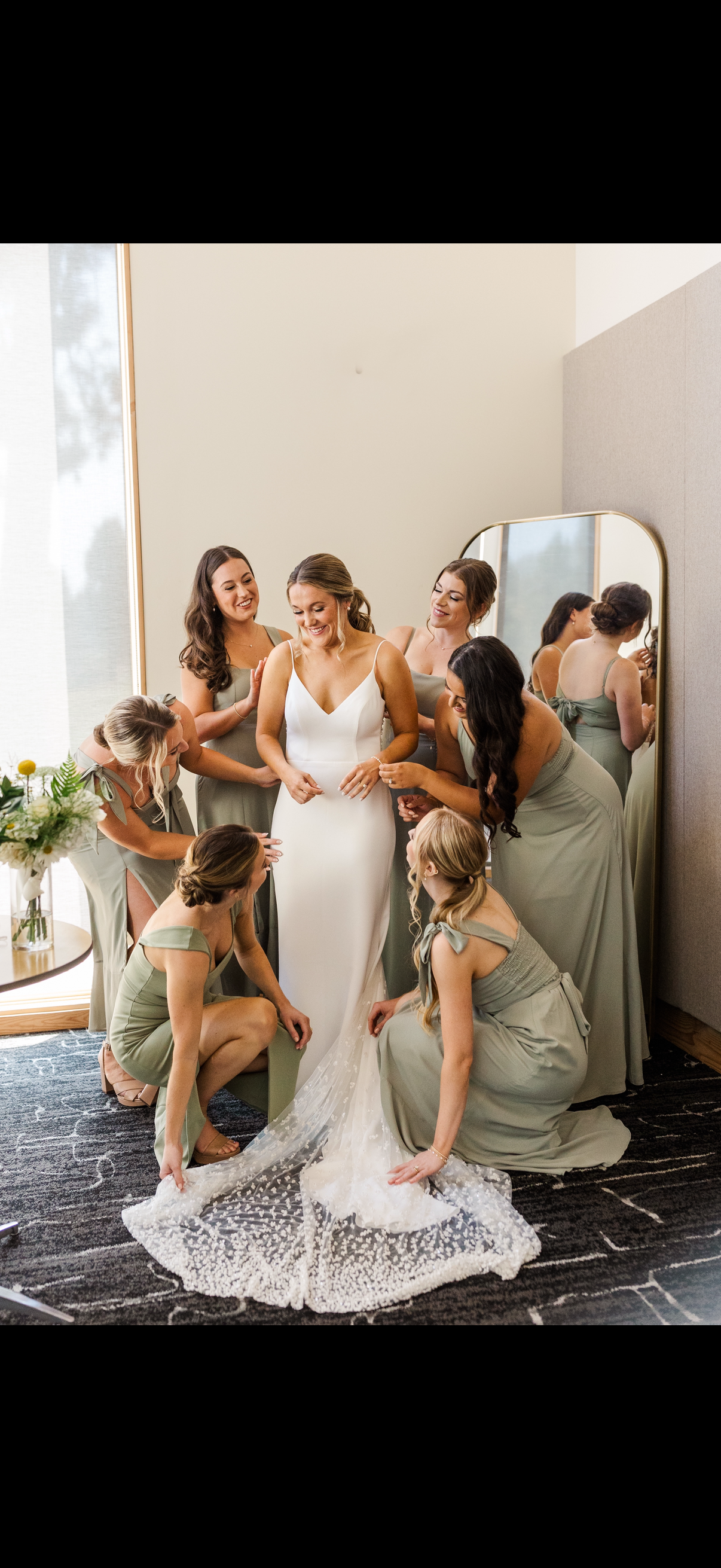 Bride with six bridesmaids around her, adjusting her white wedding dress in a room with natural light and a mirror.