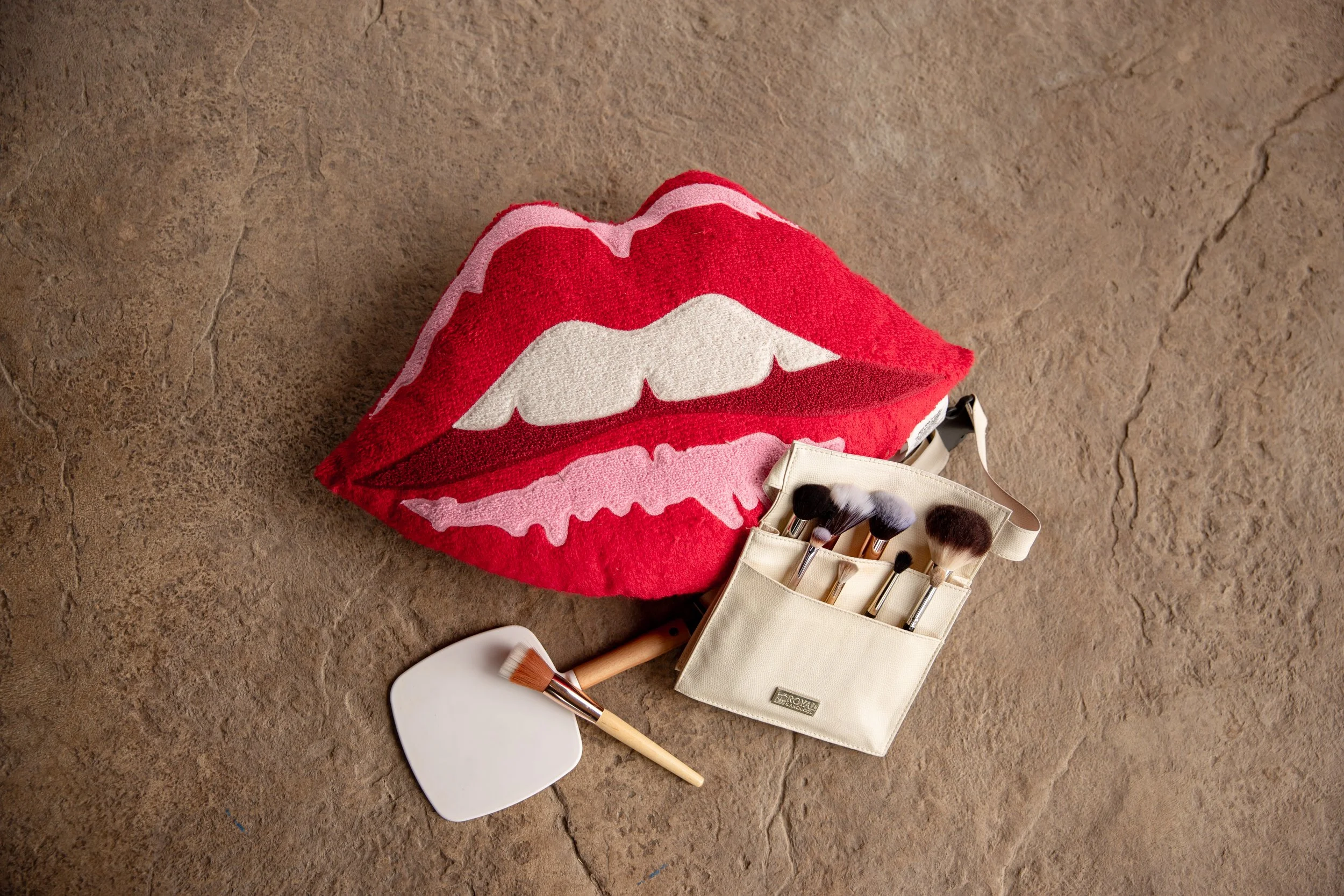 A towel with a lips and teeth design resembling a mouth with white teeth and red lips, lying on a textured brown surface next to makeup brushes, a makeup palette, and a white toiletry bag.