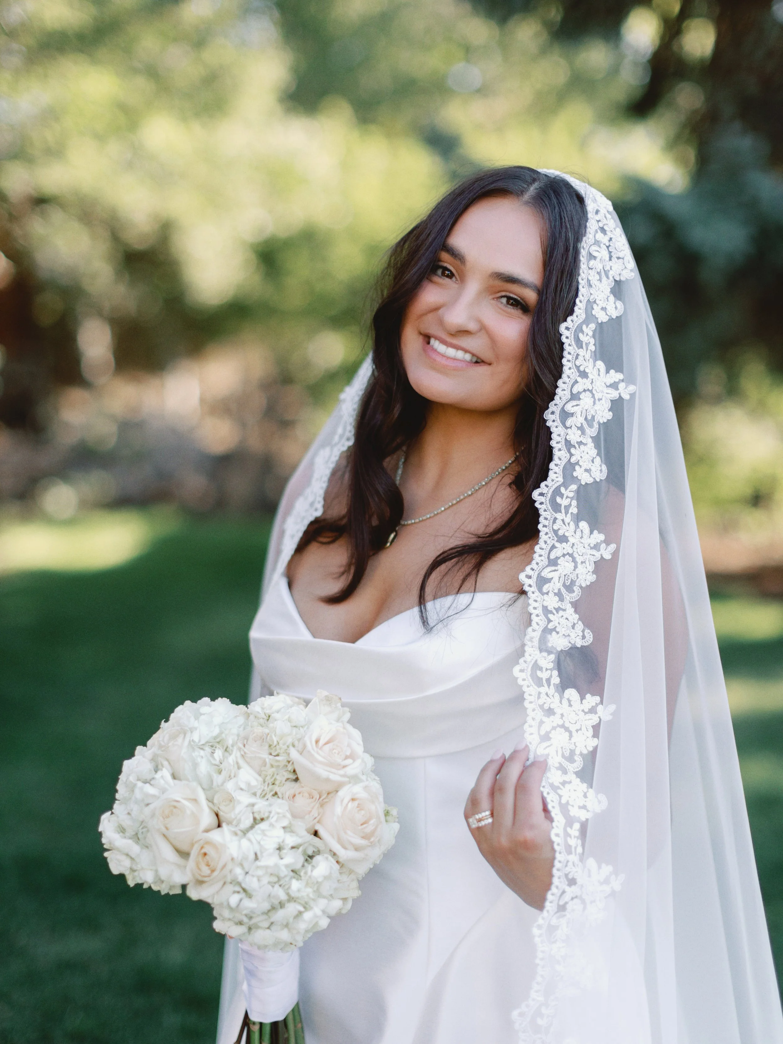 A smiling bride with dark hair, wearing a white wedding dress and veil, holding a bouquet of white flowers, standing outdoors on a green lawn with trees in the background.