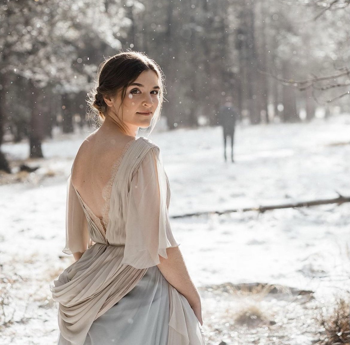 A woman in a light dress standing outdoors in a wintery landscape with snow and trees.