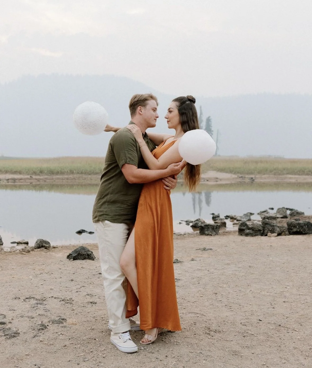A romantic couple embraces near a lake with mountains in the background, holding white paper lanterns, during daytime.