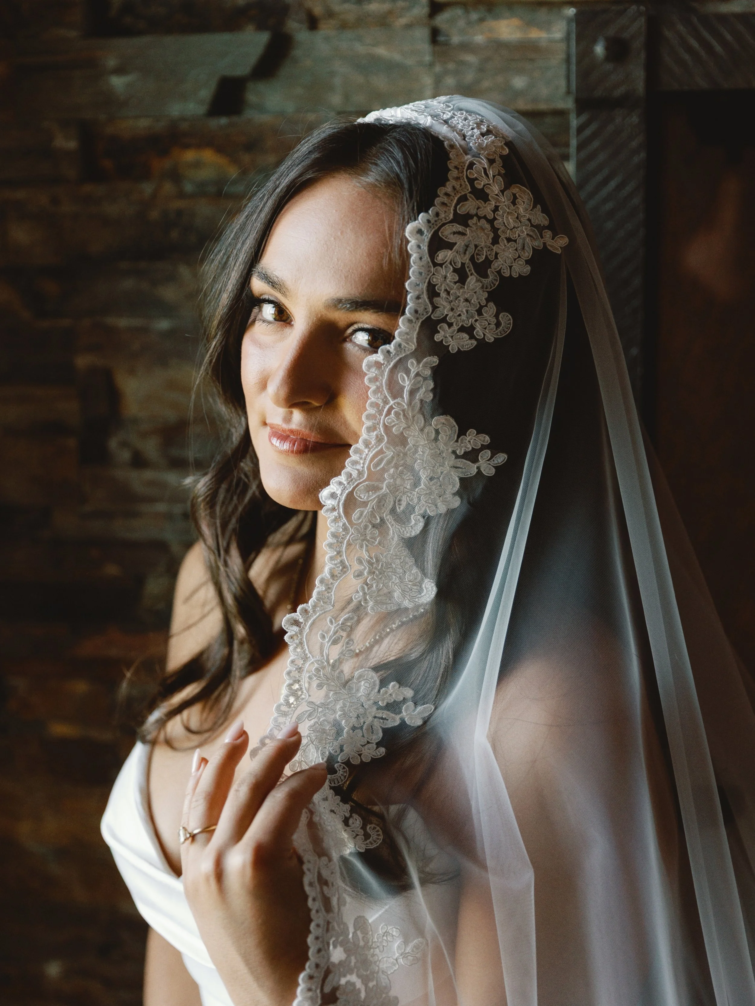 A woman with long dark hair, wearing a white dress and lace veil, touching her shoulder and looking into the camera, with a dark wooden background.