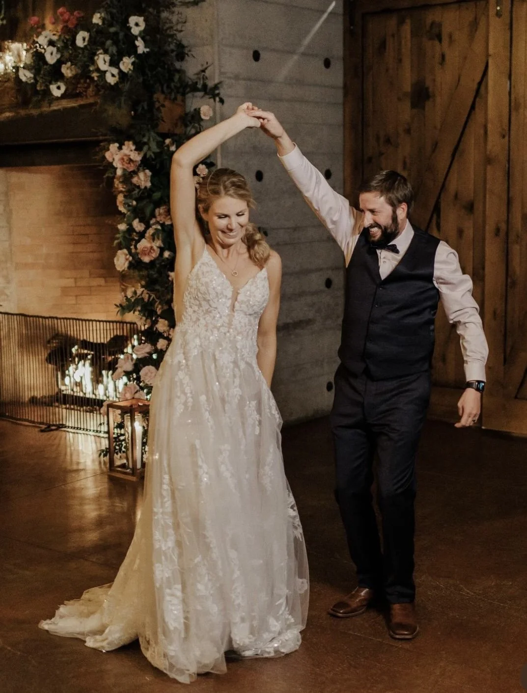 A bride and groom are dancing at their wedding celebration. The bride is wearing a white, floral lace wedding gown, and the groom is dressed in a black vest, white shirt, and bow tie. They are holding hands in a spin, smiling, with a decorated floral