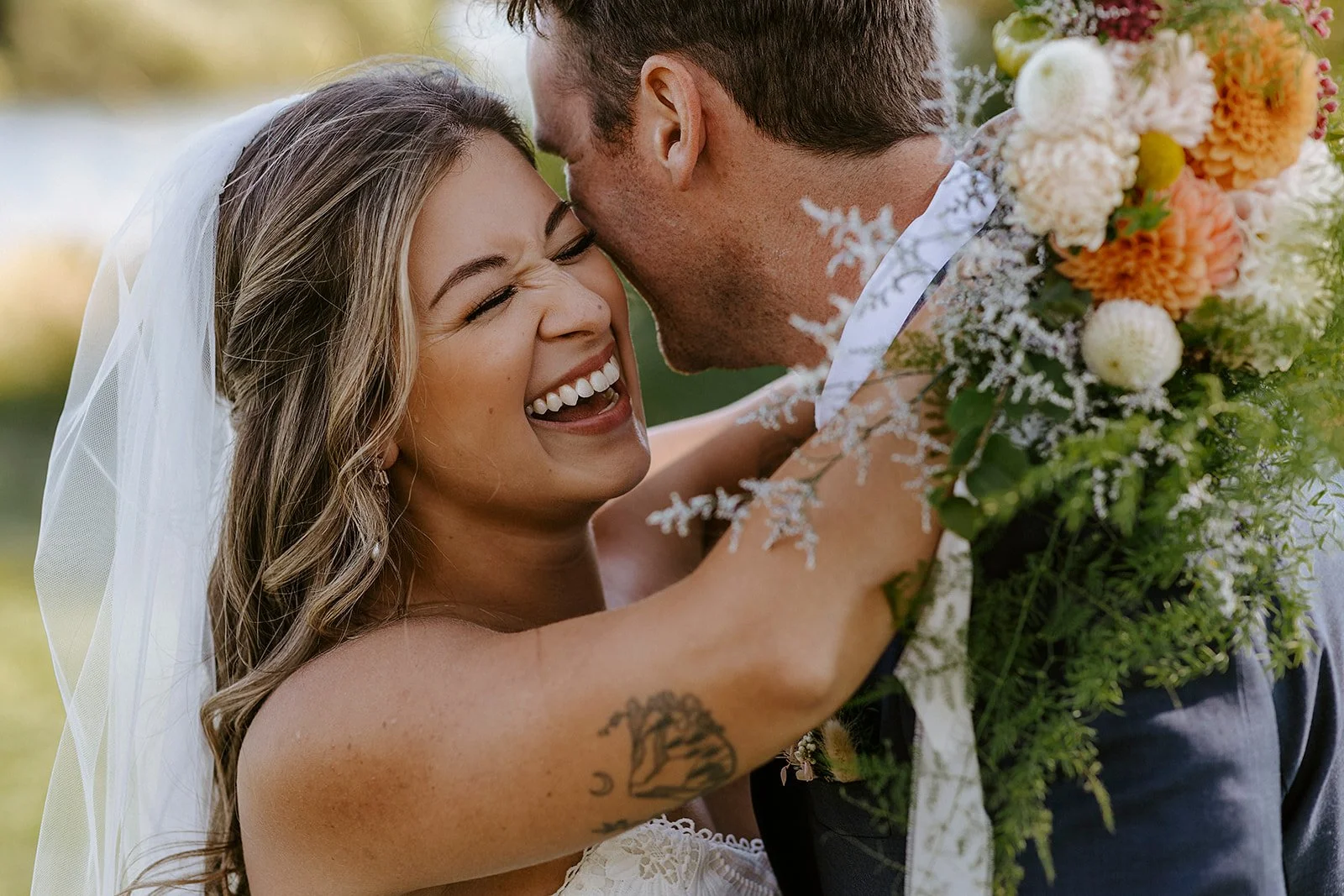 A joyful bride with long hair, wearing a wedding dress and veil, hugging a groom with a beard, who is holding a bouquet of flowers, on a sunny day outdoors.