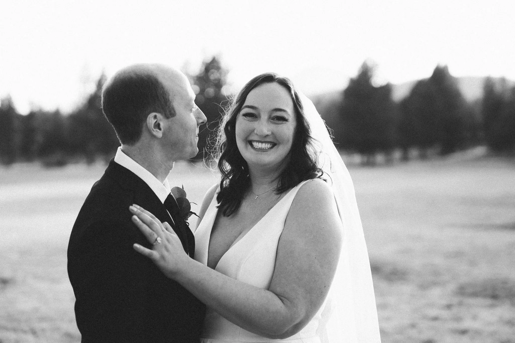 Black and white photo of a smiling bride and groom holding each other outdoors during sunset or sunrise, with trees in the background.