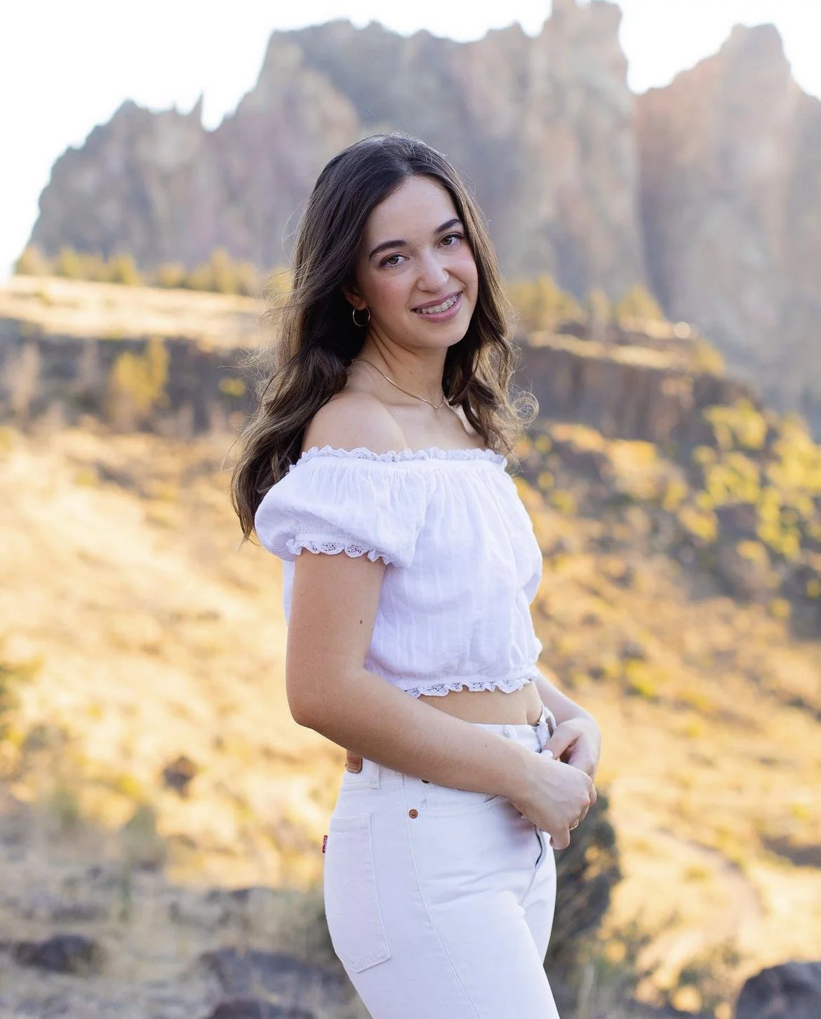 Young woman in white off-the-shoulder top and white pants standing outdoors in front of rocky mountain landscape.