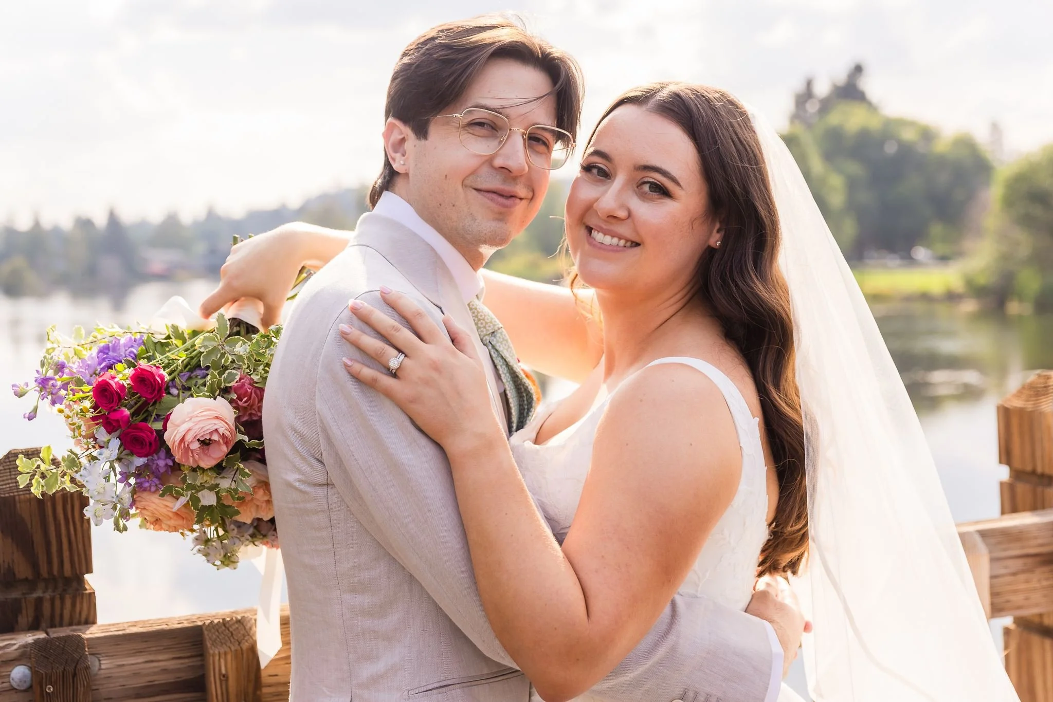A smiling bride and groom embrace outdoors during their wedding, with a bouquet of colorful flowers in the background on a wooden fence, near a body of water and greenery.