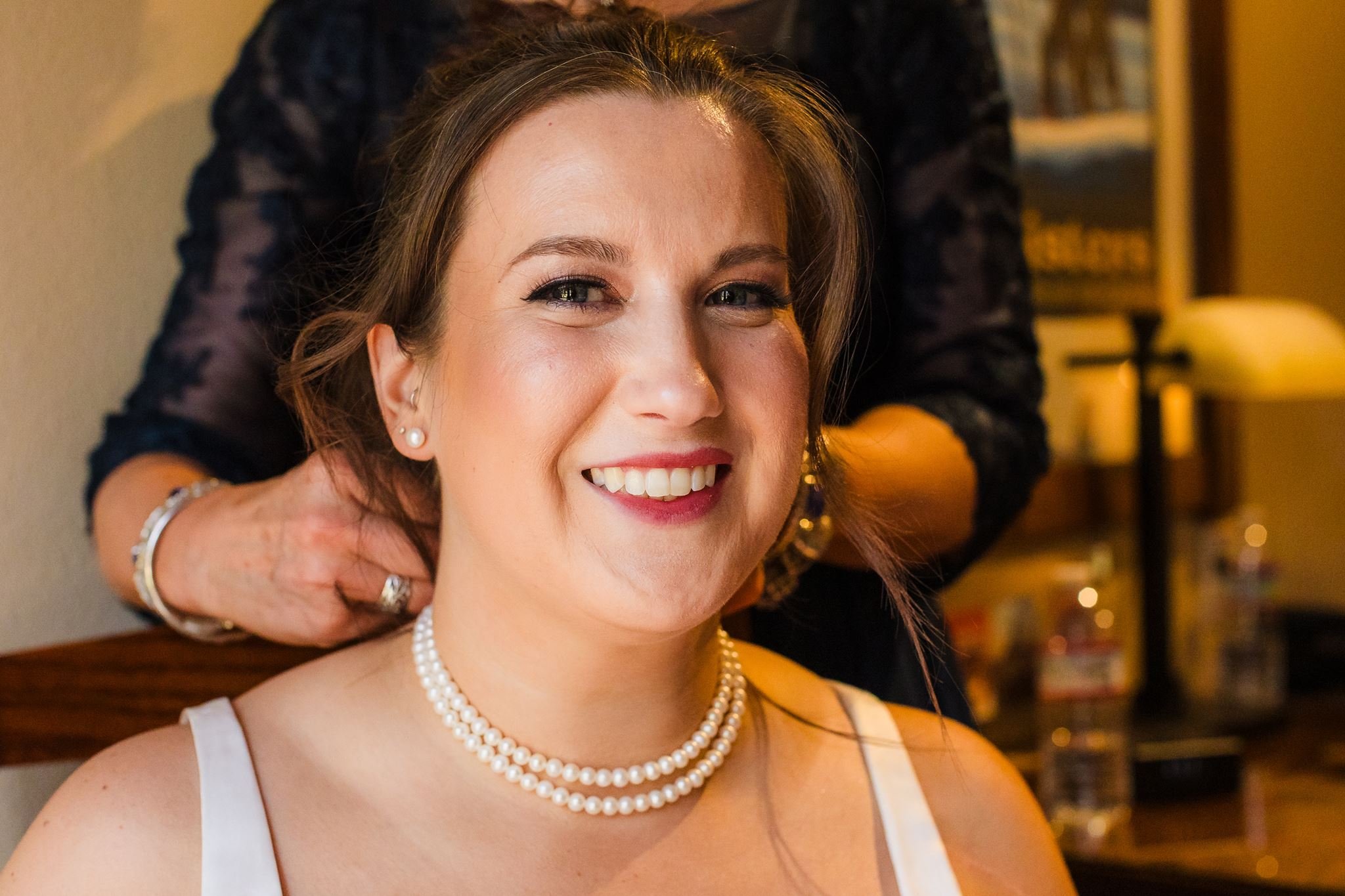 A smiling woman wearing pearl jewelry and a white top, with a person behind her adjusting her necklace, in an indoor setting with a blurred bookshelf in the background.