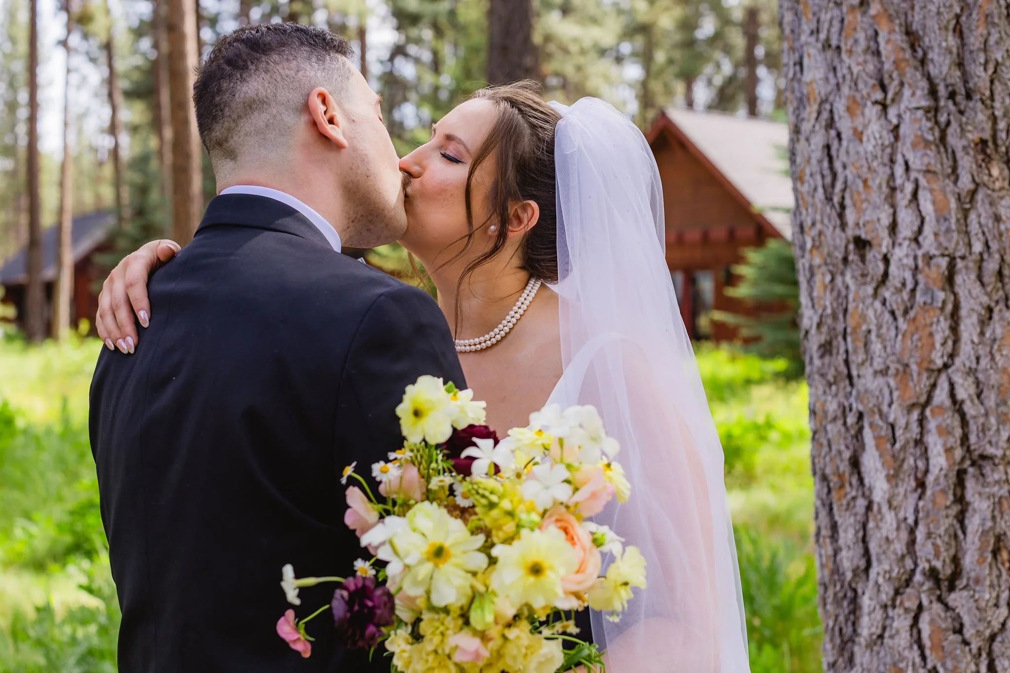 A bride and groom kissing outdoors in a forested area during their wedding. The bride is wearing a white veil and pearl necklace, holding a bouquet of yellow and white flowers. The groom is in a black suit with a white shirt. There are tall pine tree