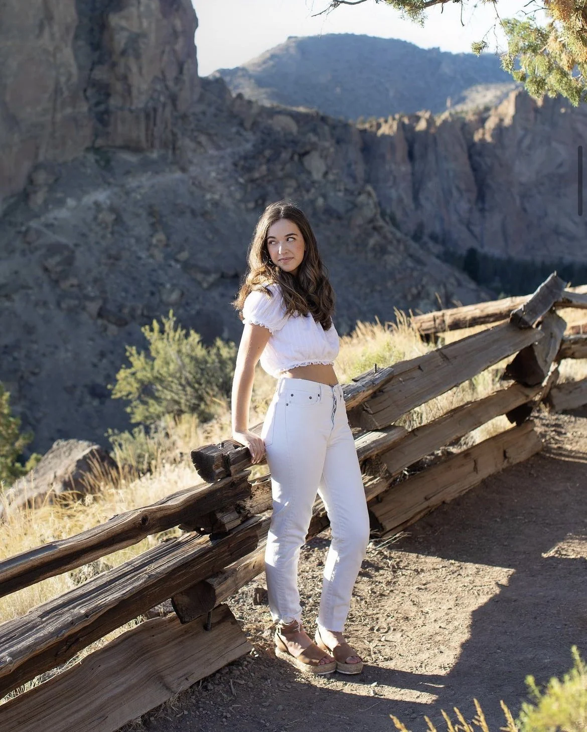 A woman standing outdoors by a rustic wooden fence with rocky mountainous terrain in the background, wearing a white crop top, white pants, and sandals.