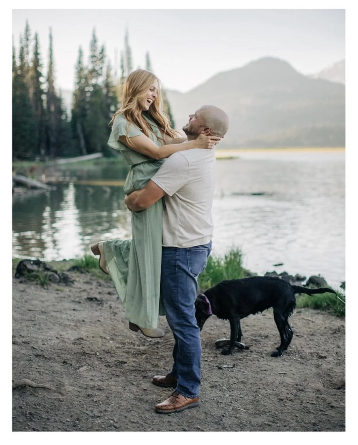 A man lifting a woman in a green dress by a lakeside, with a black dog nearby and mountains in the background.