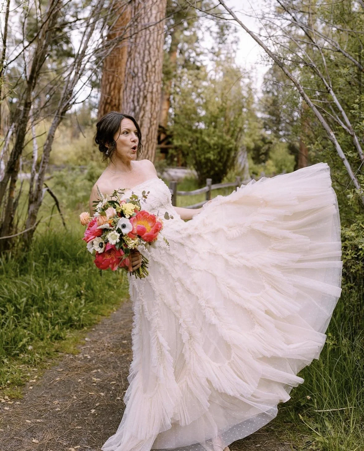 A woman in a white wedding dress holding a colorful bouquet of flowers, standing outdoors on a dirt path surrounded by trees and greenery.