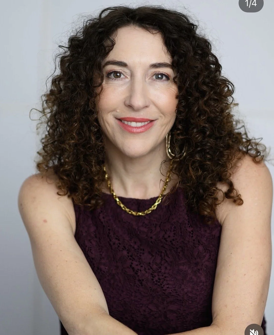 A woman with curly brown hair smiles at the camera, wearing a purple lace top, gold necklace, and matching earrings.