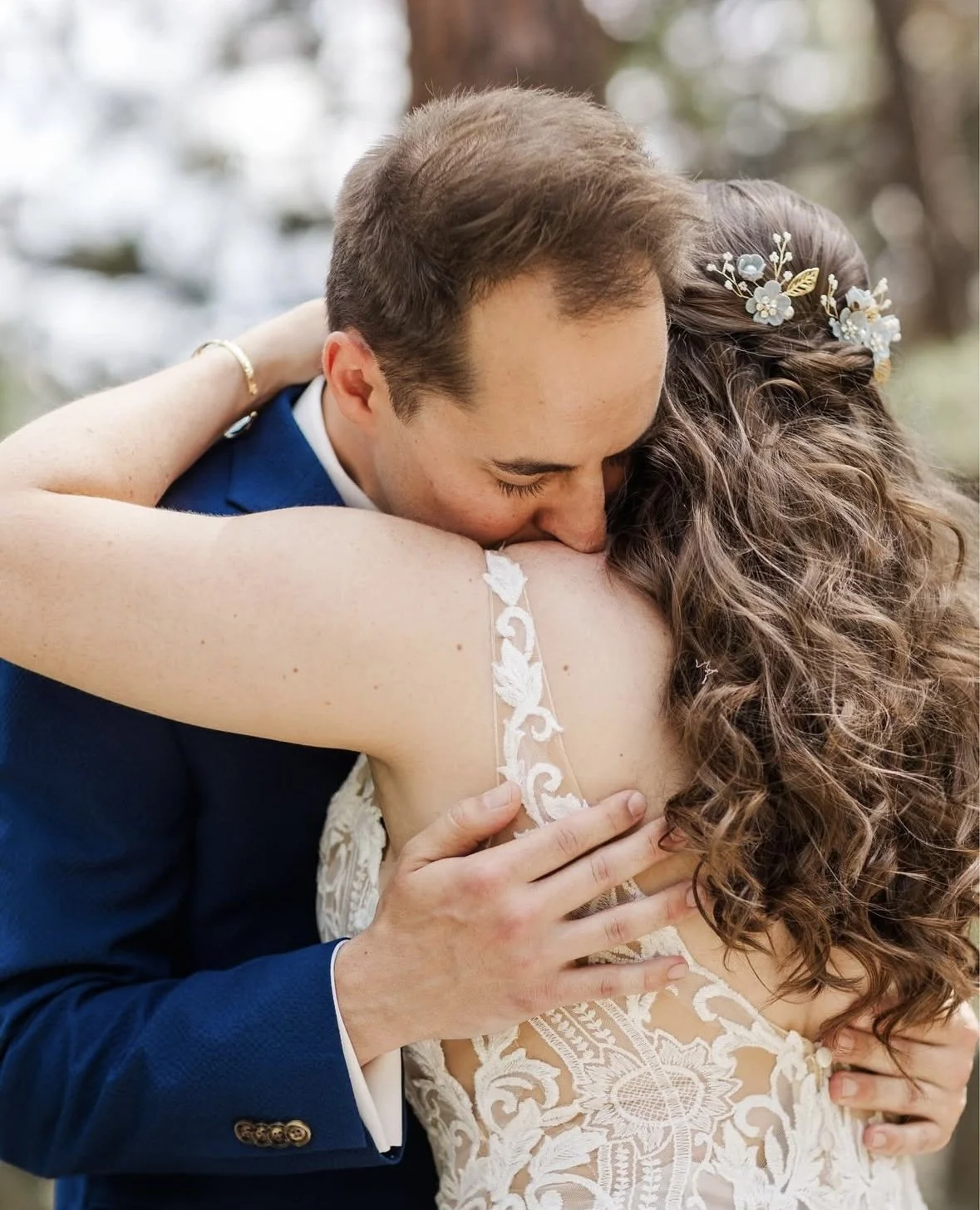 A man and woman embracing in a wedding hug outdoors, with the man wearing a blue suit and the woman in a lace wedding dress, with floral hair accessories.