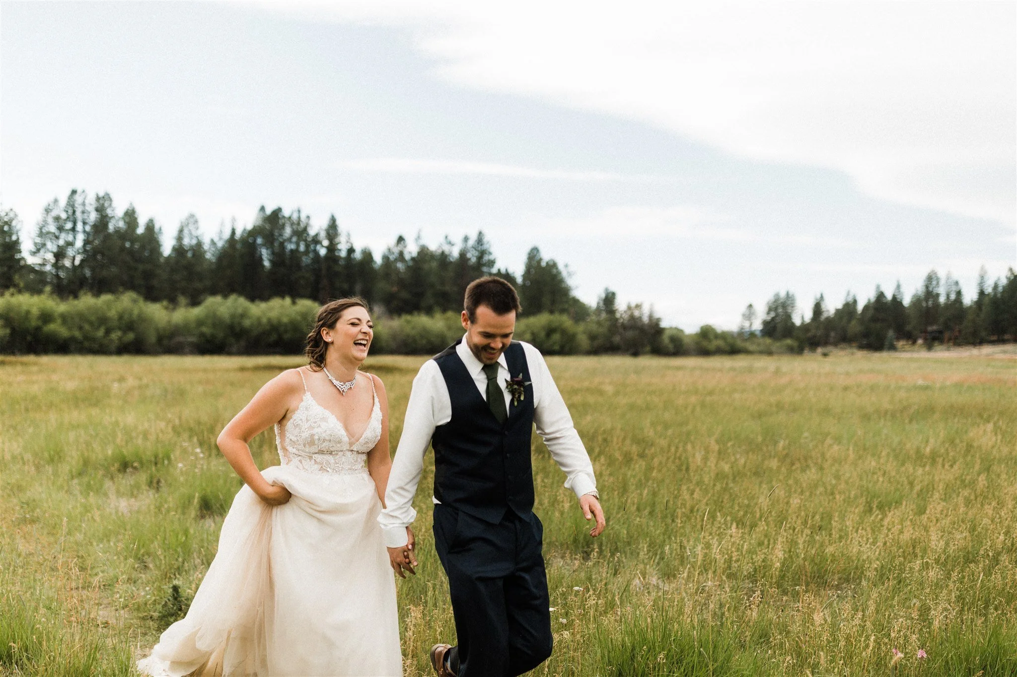 A smiling bride and groom walking hand-in-hand across a grassy field outdoors on a cloudy day, with trees in the background.