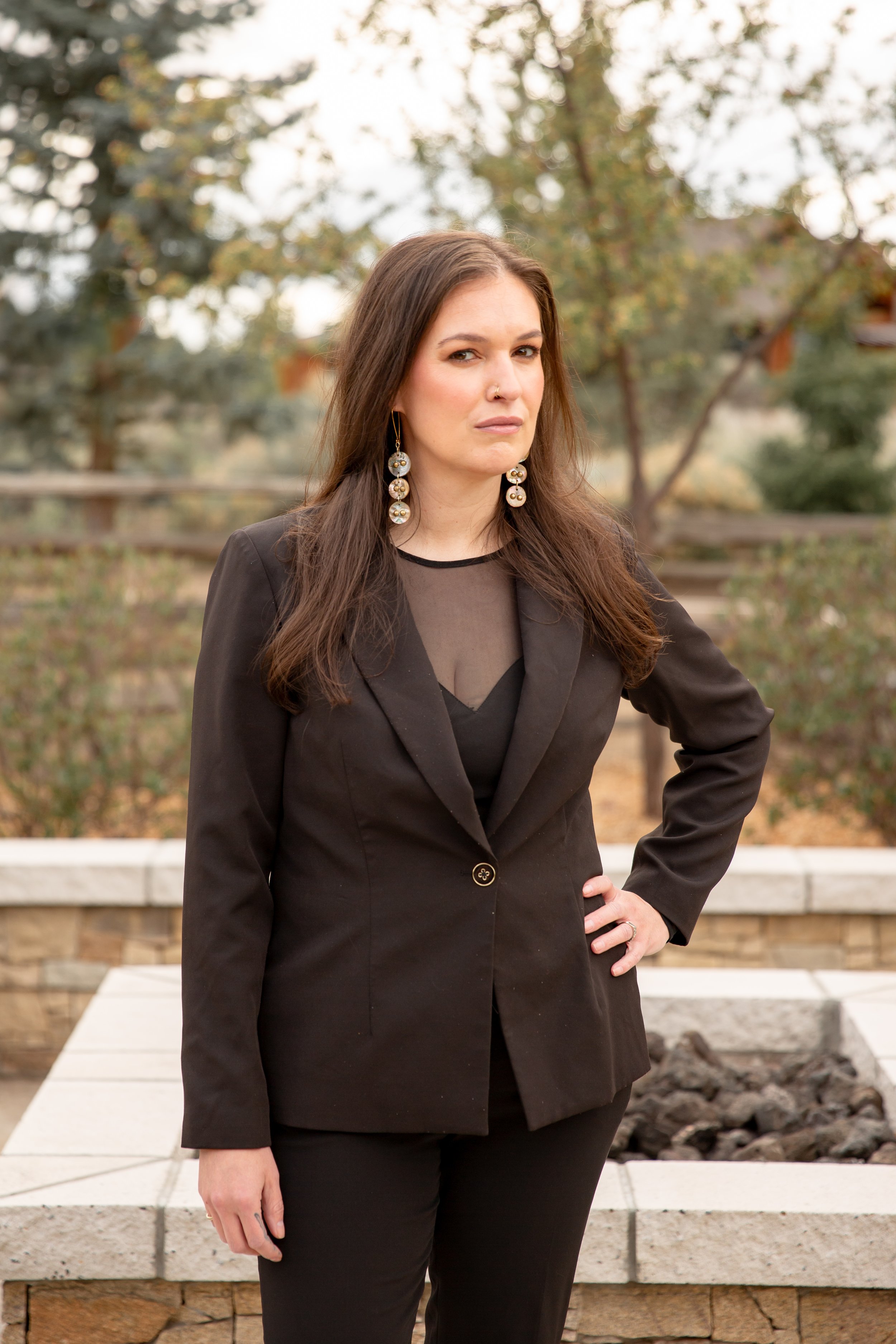 A woman with long brown hair, wearing a black blazer with a single button and black pants, stands outdoors with a neutral expression. She has large dangling earrings and a nose ring, with her right hand on her hip, and is in front of a landscaped background with trees and a low stone wall.