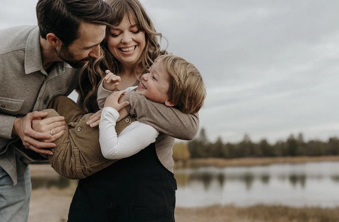 A family of three enjoying a moment outdoors near a lake, with the father and mother smiling and holding their young son, who is smiling and playfully tugging at the mother's arm.