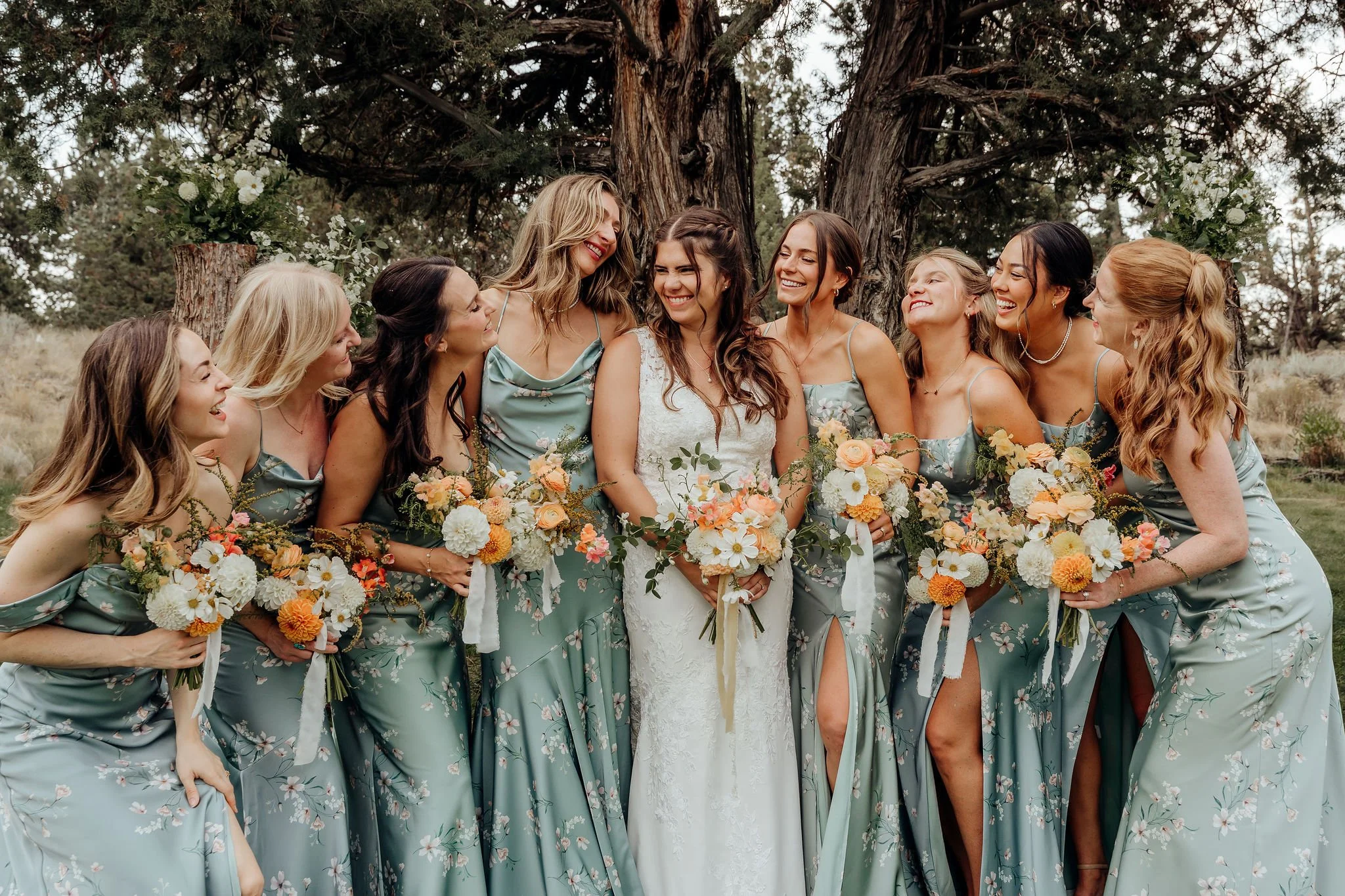 A group of eight women dressed in matching blue floral dresses, gathered outdoors around a bride in a white wedding gown, smiling and laughing in front of a large tree.