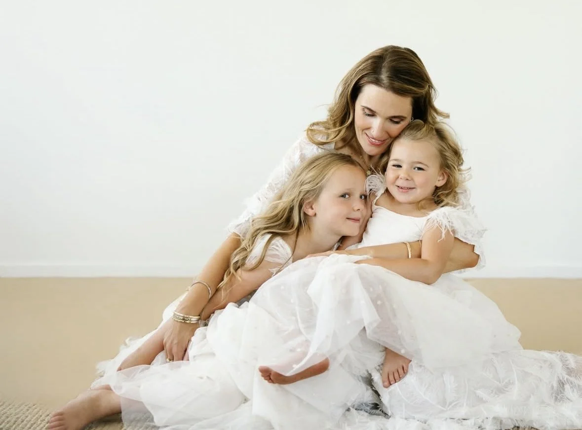A woman and three young girls dressed in white, sitting on the floor and hugging each other in a minimalistic room.