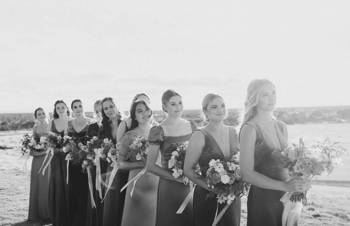 A line of women in formal dresses holding bouquets of flowers, standing outdoors near water on a sunny day.