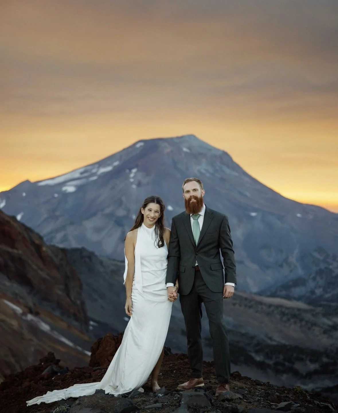 A newlywed couple holding hands and smiling outdoors at sunset, with a mountain in the background.