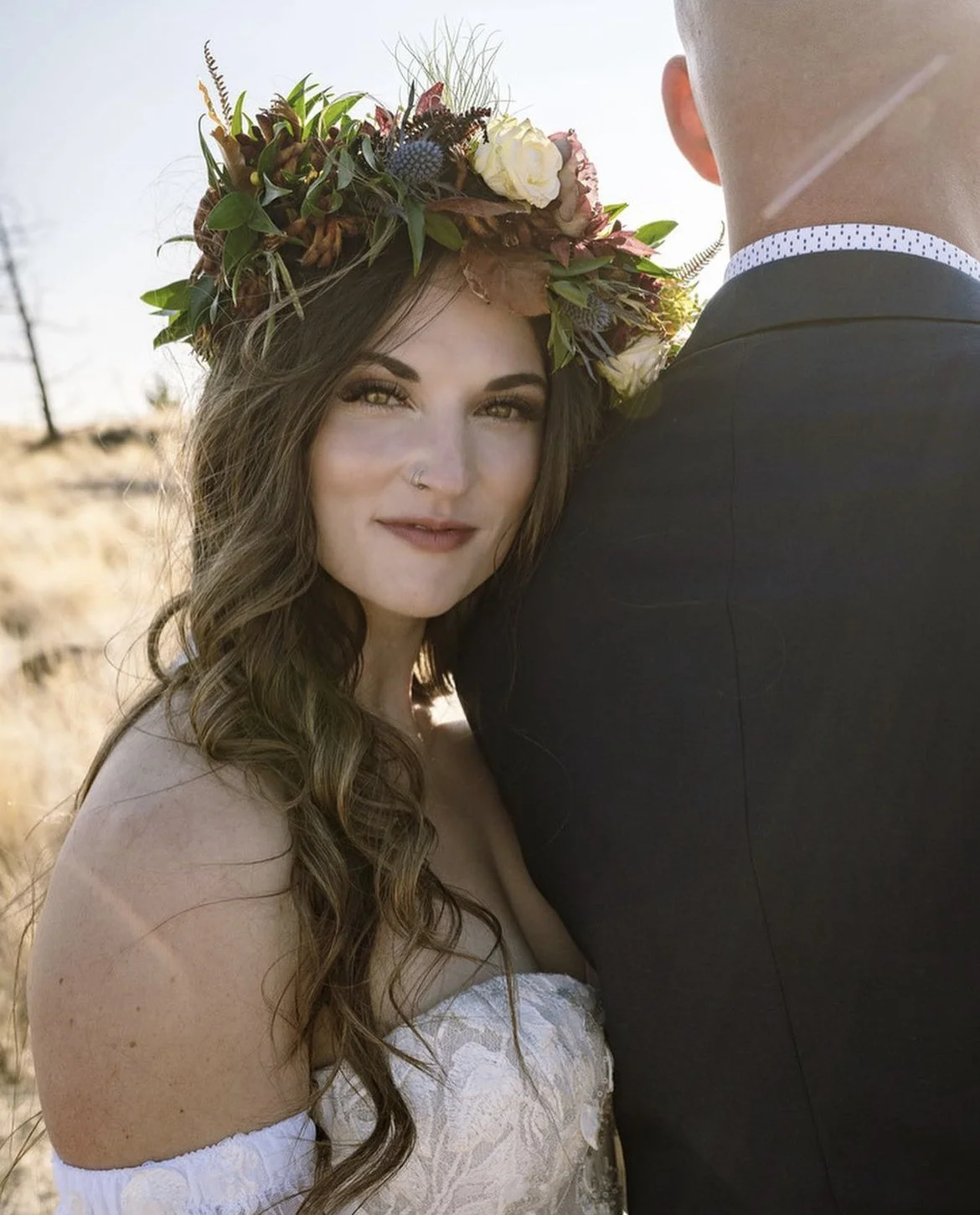 A woman with long wavy brown hair wearing a white off-the-shoulder dress and a colorful flower crown, standing next to a man in a black suit outdoors.