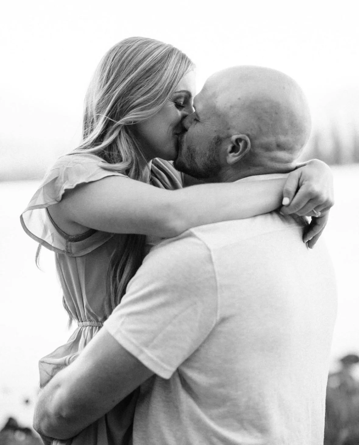 A woman and a man embrace and kiss outdoors, with the woman wearing a dress and the man with a shaved head, in a black-and-white photo.