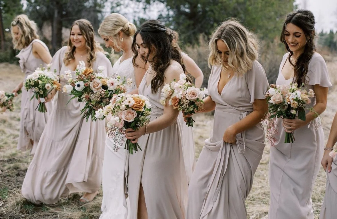 A group of women in light-colored dresses holding bouquets walking outdoors on grassland with trees in the background.