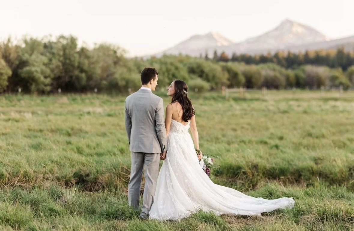 A bride and groom holding hands in a field with mountains in the background.
