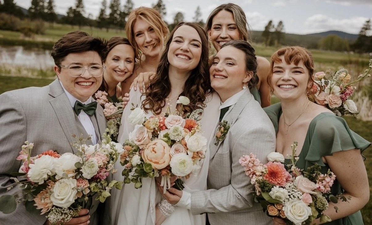 A group of seven smiling people, including a bride and groom, posing outdoors with a lake and trees in the background, holding bouquets of flowers during a wedding celebration.