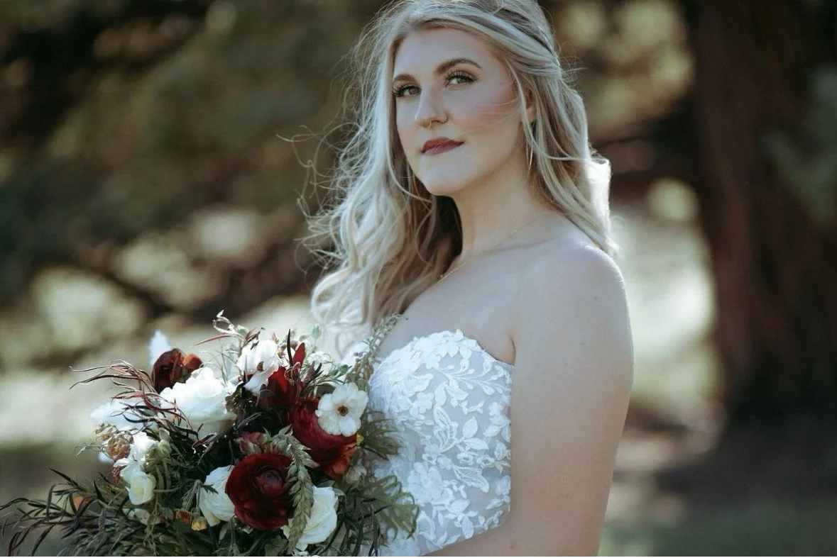A bride with long, wavy blonde hair wearing a white strapless wedding gown holding a bouquet of red, white, and green flowers outdoors in front of blurred trees.