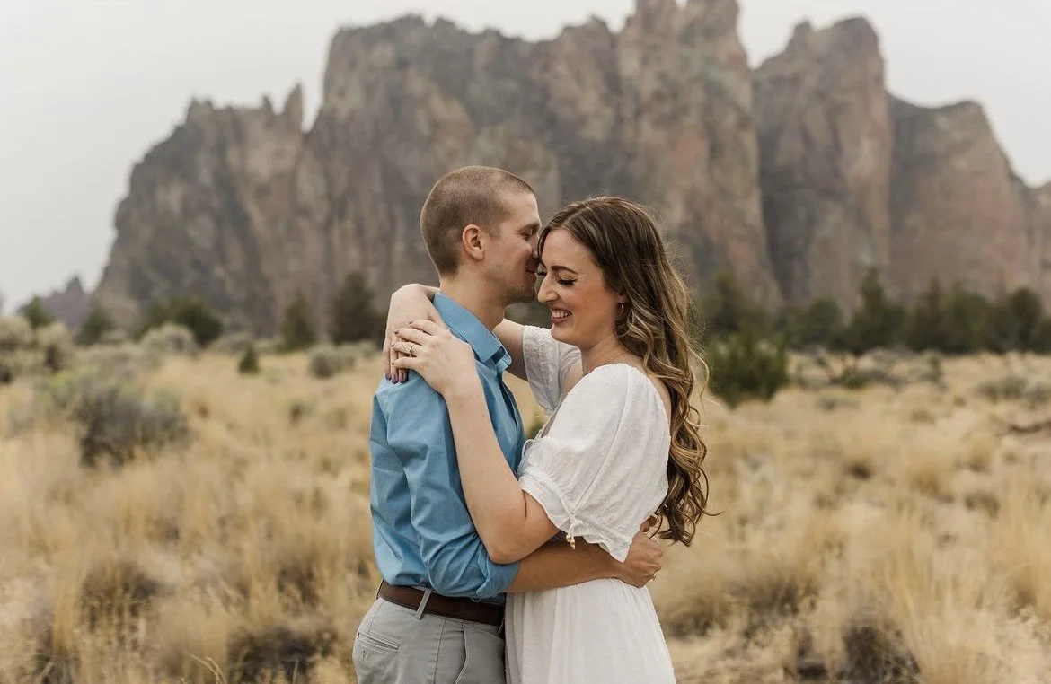 A couple embraces and smiles in a field with mountains in the background during daytime.