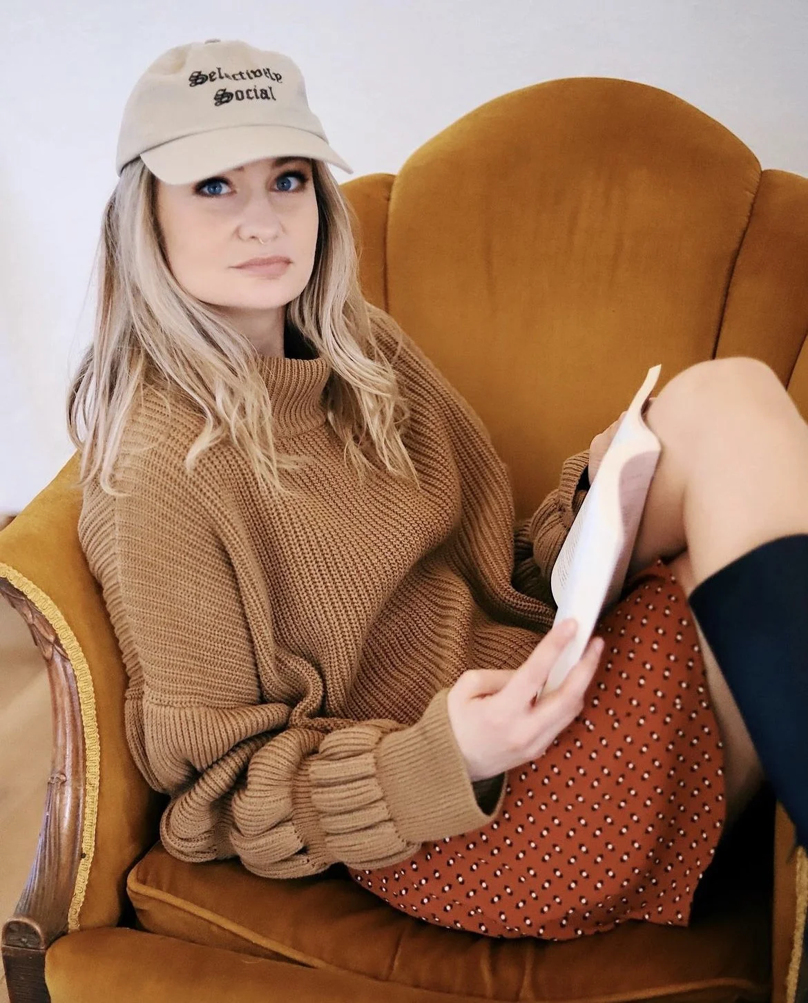 A woman with blonde hair and blue eyes, wearing a beige baseball cap with the words 'Selective Social', a brown knit sweater, and a polka dot skirt, sitting on an orange velvet armchair with wooden armrests, holding a piece of paper. The background i