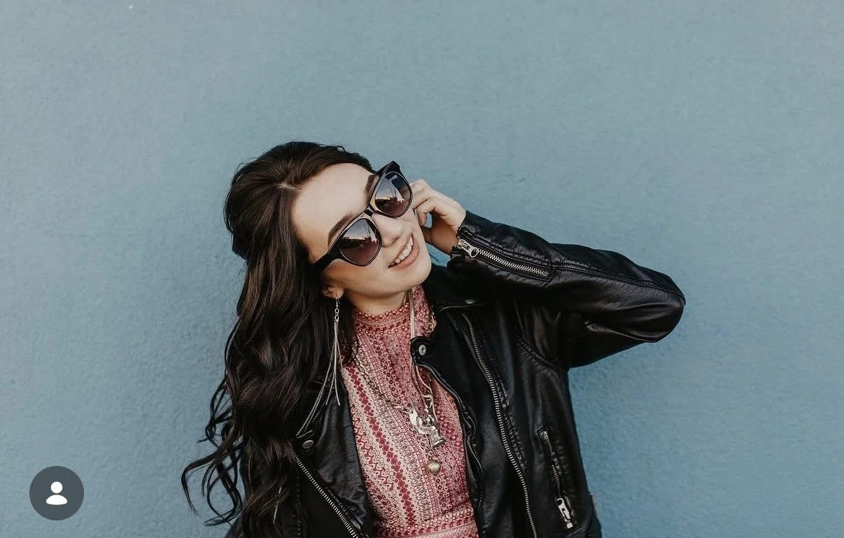 A woman with long dark hair, wearing dark sunglasses, a black leather jacket, and a pink patterned top, is smiling and posing against a blue wall.