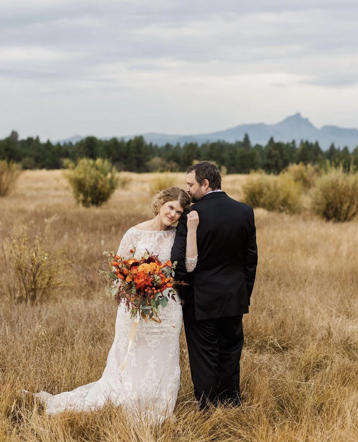 A bride and groom standing in a field with tall grass and shrubs, with mountains and a cloudy sky in the background. The bride is holding a bouquet of orange and red flowers and is leaning on the groom, who is wearing a black suit.