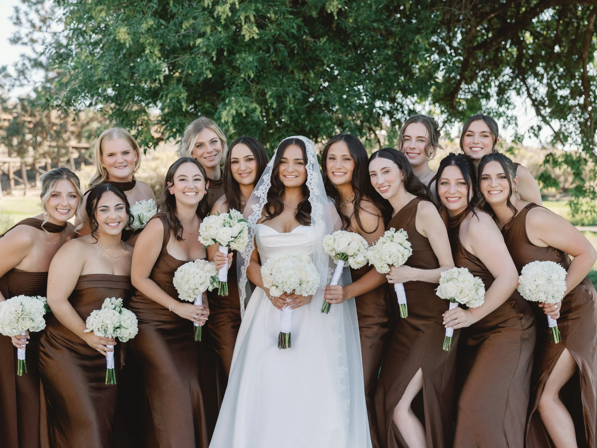 Group of women, one in a white wedding dress and veil, surrounded by bridesmaids in brown dresses, holding white flower bouquets outdoors under a large leafy tree.