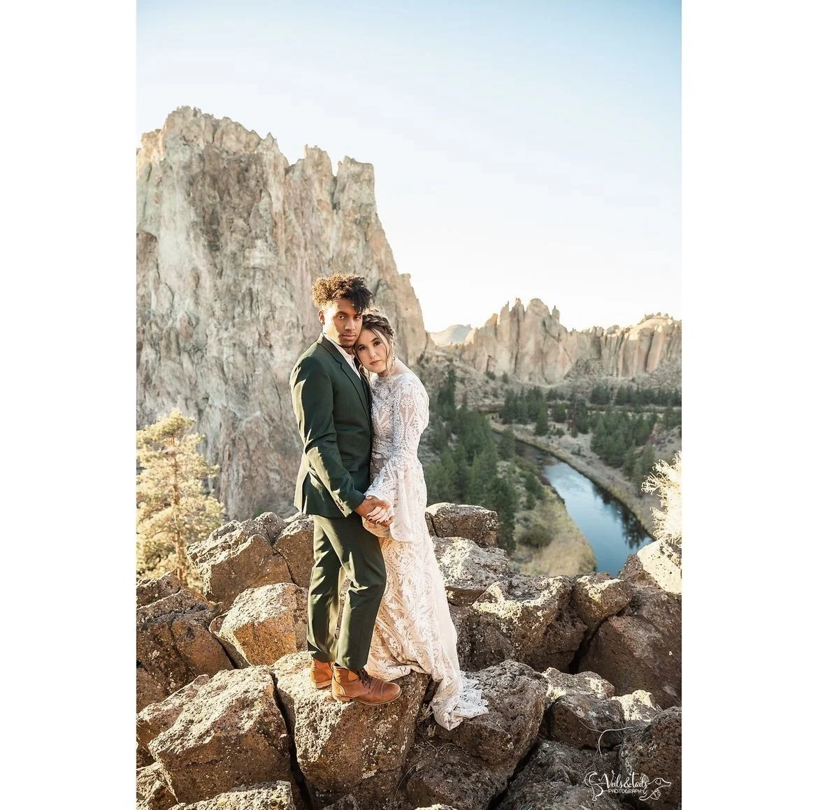 A couple in wedding attire standing on rocks with a scenic mountainous landscape and river in the background during sunset.