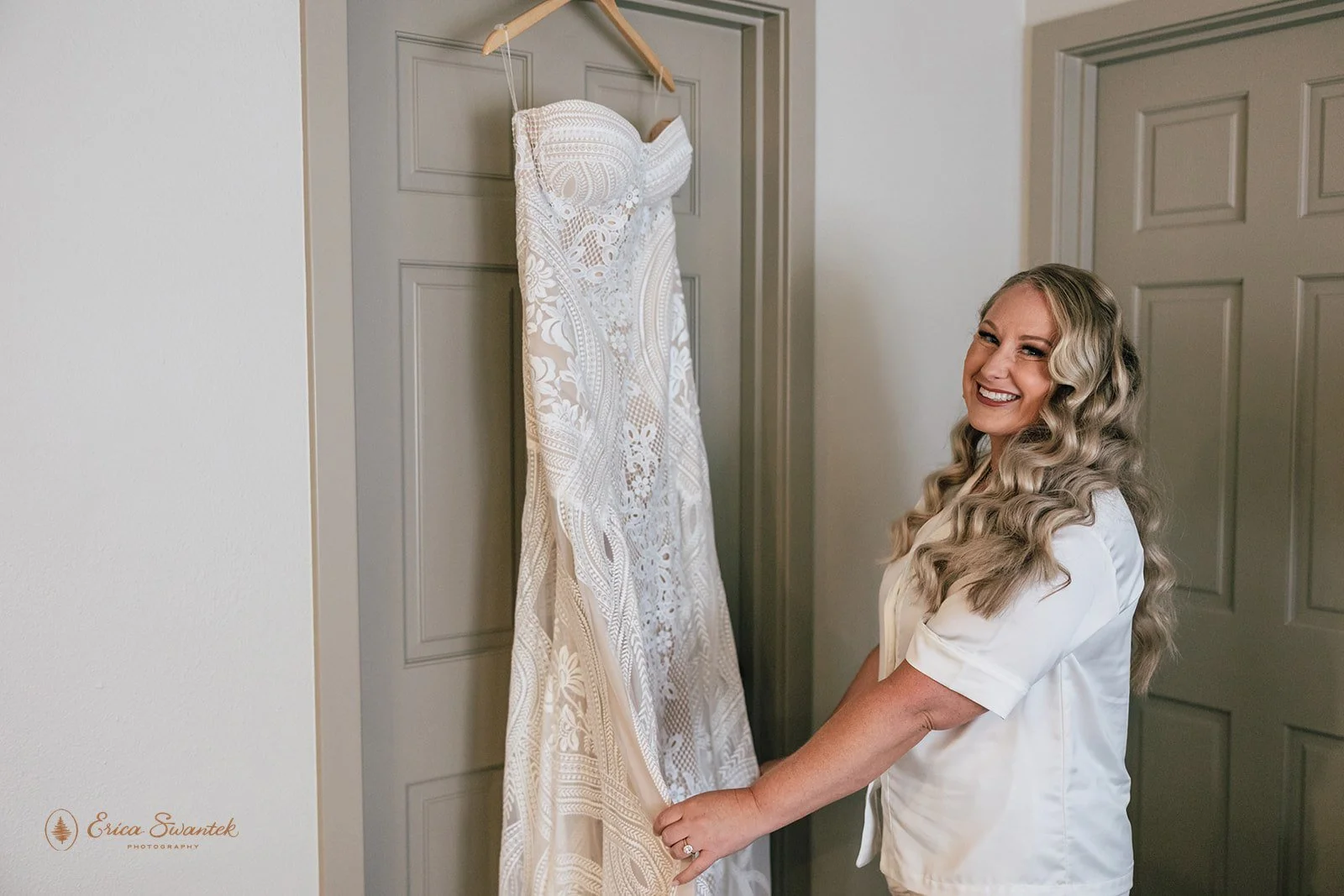 A woman with long, wavy blonde hair smiling as she holds a wedding dress hanging on a door.