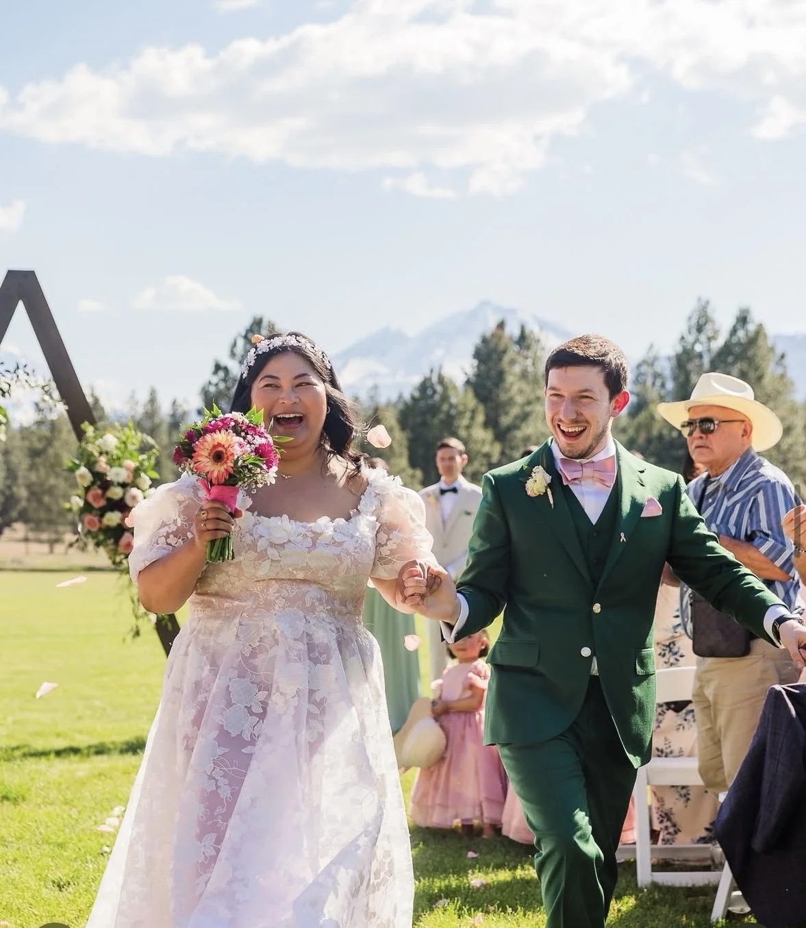 Happy bride and groom walking outdoors at a wedding ceremony, smiling and holding hands, with guests in the background and a scenic mountain landscape.