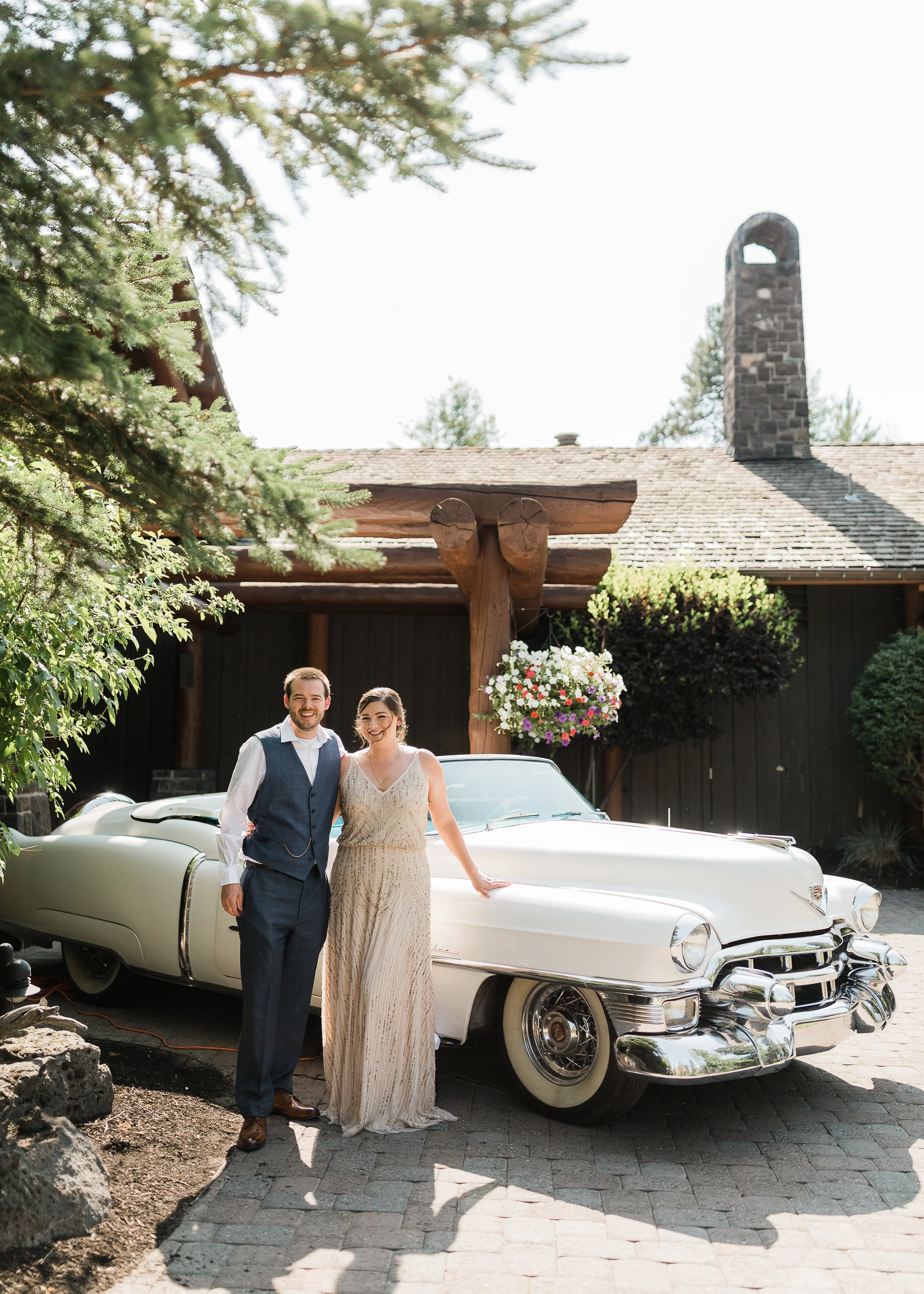 A man and woman in formal attire standing together in front of a vintage white Cadillac car, with a large wooden building with a stone chimney and hanging flower basket in the background, amidst greenery and rocks.