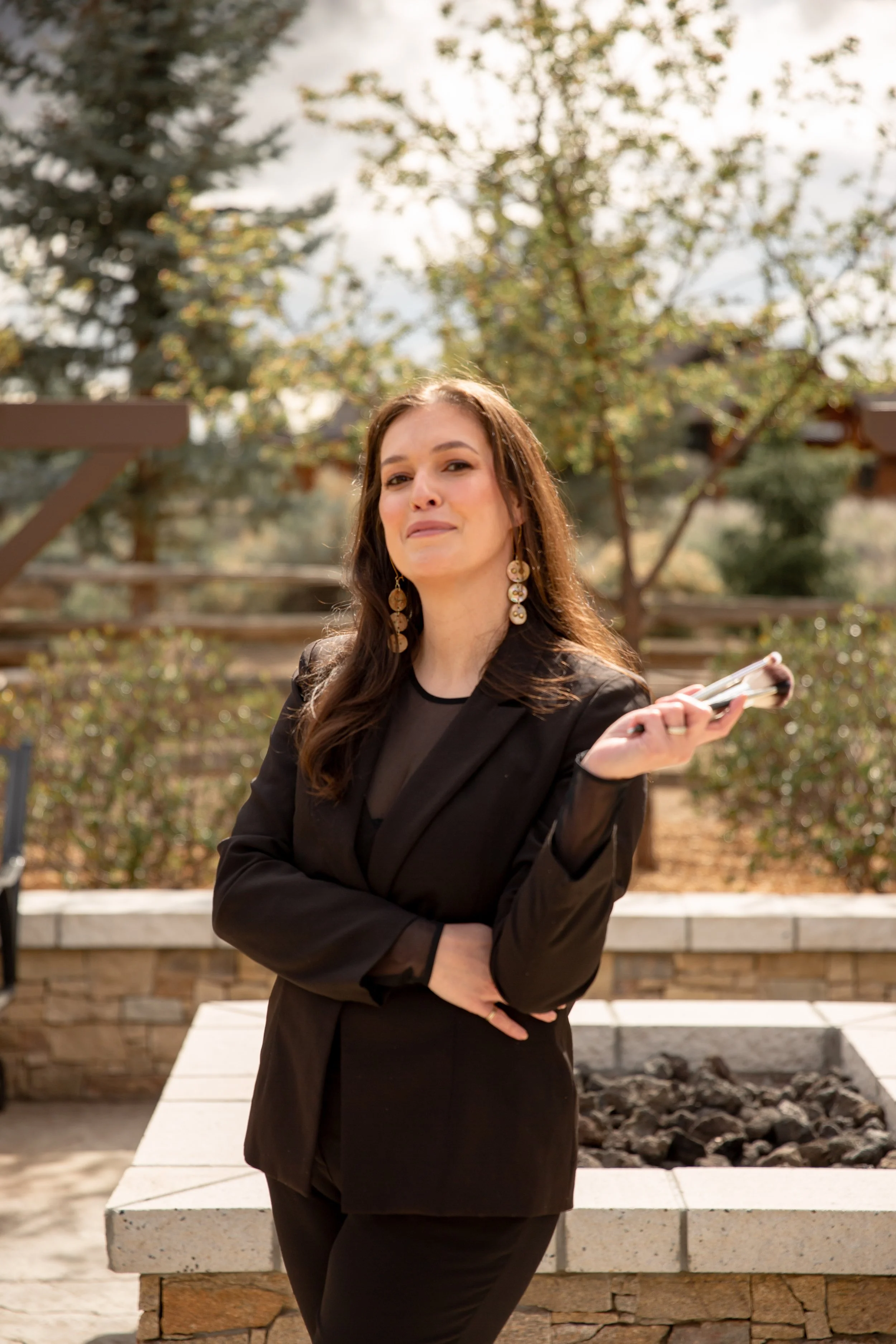 A woman with long dark hair, wearing a black blazer and earrings, stands outdoors in front of a low stone wall and plants, holding a smartphone, with a large tree and a cloudy sky in the background.
